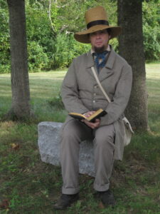 Man dressed in 19th century clothing aand aa straw hatt sittting on a rock holding a book.